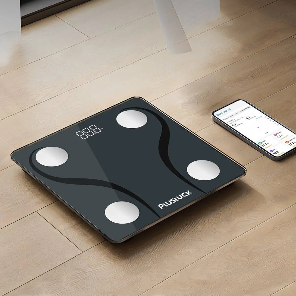 Black digital body fat scale on a wooden floor with a smartphone displaying app.